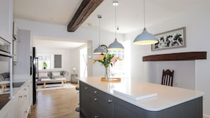 A modern kitchen with a central island, pendant lights, and a dining area with a chair and table. - Plas Coch (Saint Asaph)
