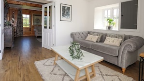 A cozy living room with a grey sofa, a white coffee table, and a wooden floor. There is a window with a view of greenery outside. - Plas Coch (Saint Asaph)