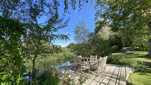 Um deck de madeira com mobiliário para área externa com vista para um lago, rodeado por vegetação exuberante e árvores.