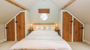 Quarto com teto inclinado, vigas de madeira, cama com colcha listrada, duas mesas de cabeceira com abajures e uma janela com vista.