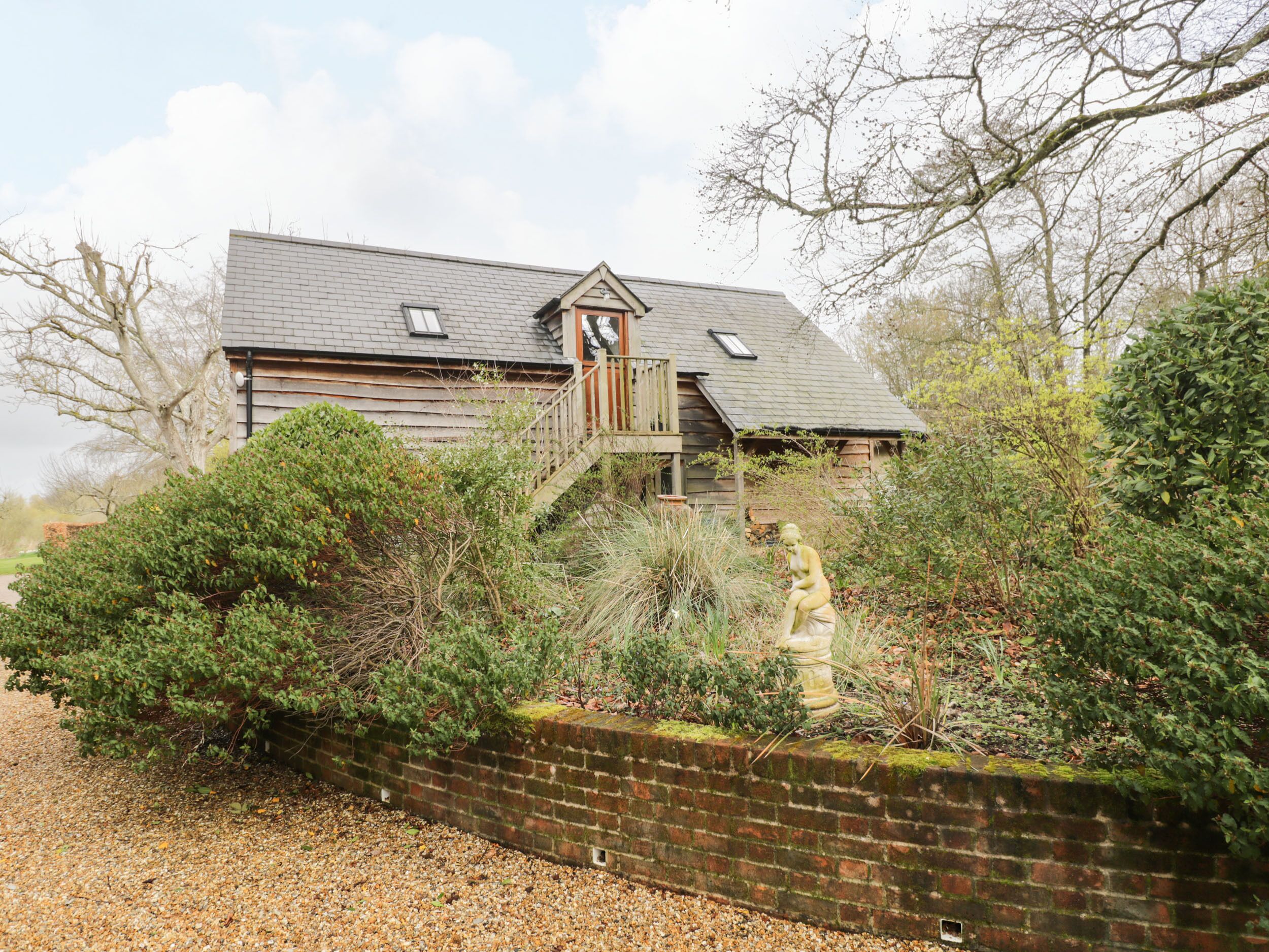 A rustic wooden house with a sloped roof, a wooden staircase leading to a deck, and a brick wall in the foreground.