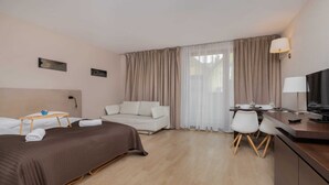 Quarto de hotel com cama, sofá, escrivaninha com cadeiras, televisão e janela com cortinas.