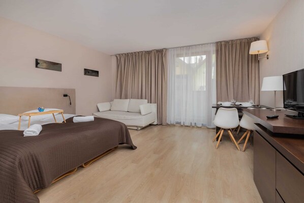 A hotel room with a bed, a sofa, a desk with chairs, a TV, and a window with curtains.