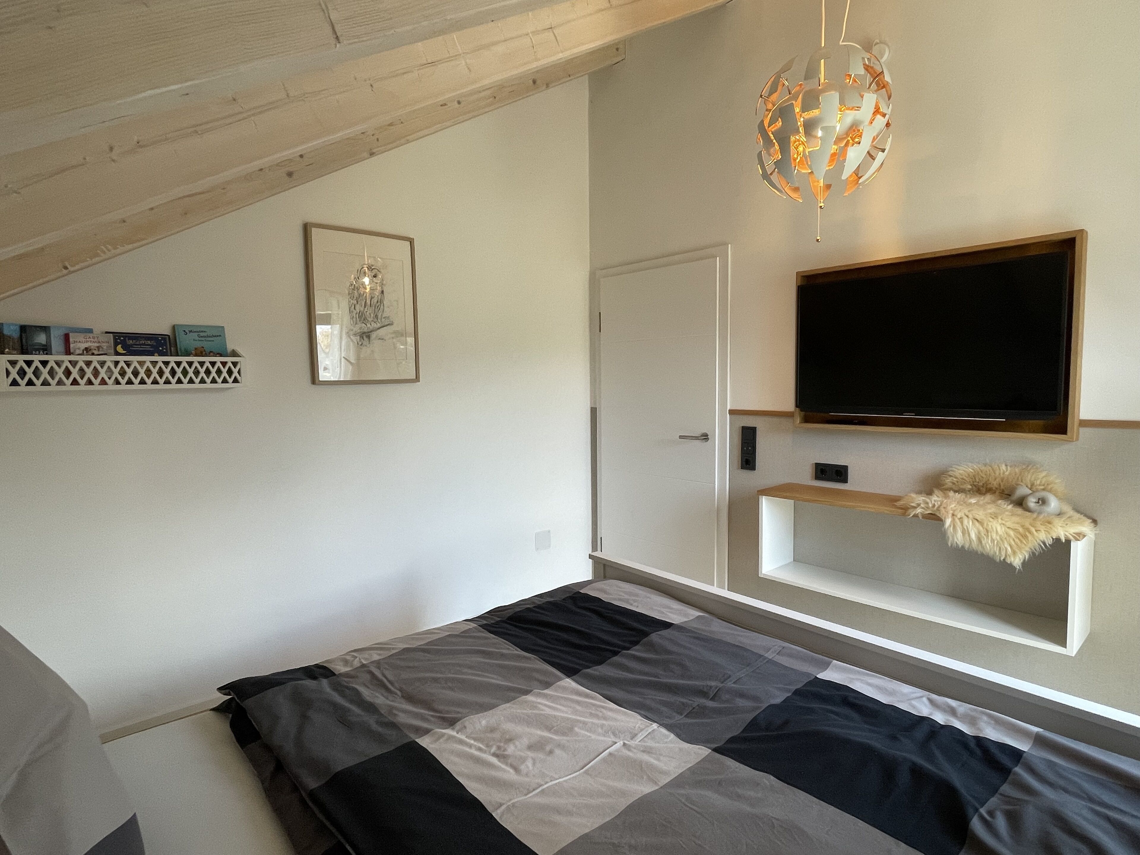Un dormitorio con una cama, un televisor montado, una estantería con adornos y un cuadro en la pared.