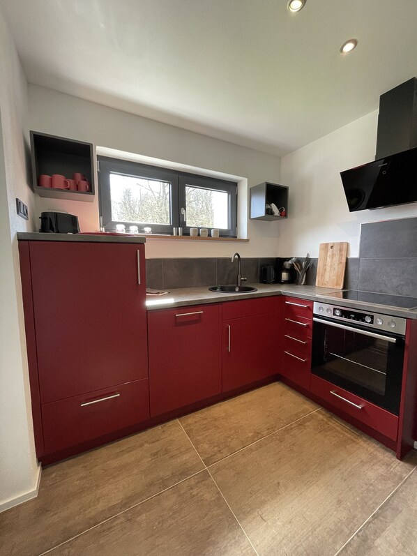 A modern kitchen with red cabinetry, a built-in oven, and a window above the sink. - Vacation home with large sun terrace (Edertal)