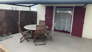 Un patio con una mesa redonda y cuatro sillas de madera, un banco de metal y una puerta corrediza de vidrio con un persiano rojo.