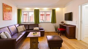 Una sala de estar con un sofá de cuero marrón, una mesa de centro de madera y un banco rojo. Hay un televisor montado en la pared y una ventana con cortinas verdes.