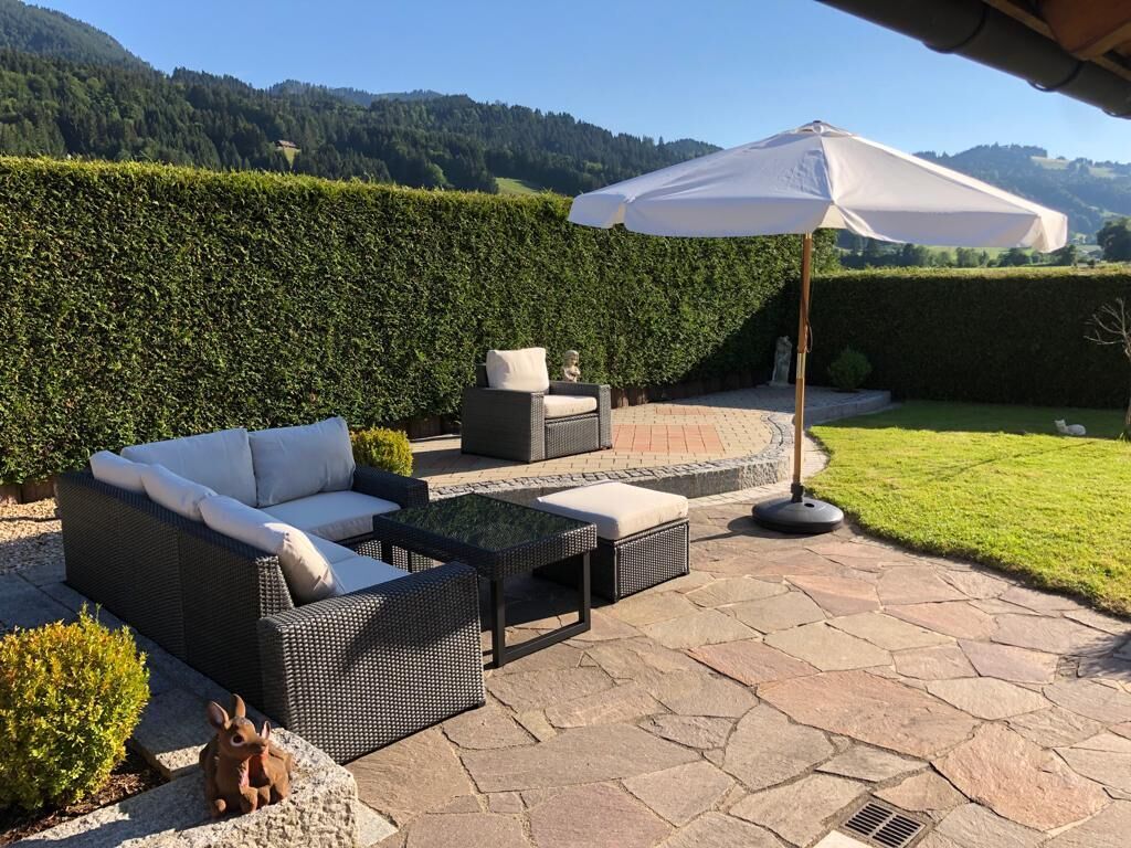 An outdoor patio with wicker furniture, a white umbrella, and a view of a green landscape.