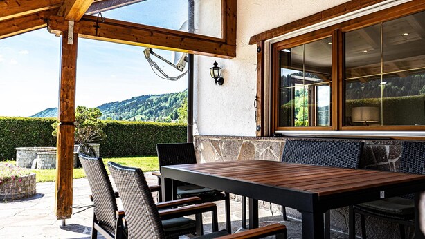 Eine Terrasse im Freien mit Holztisch und -stühlen, einer Steinmauer und Blick auf eine Berglandschaft.