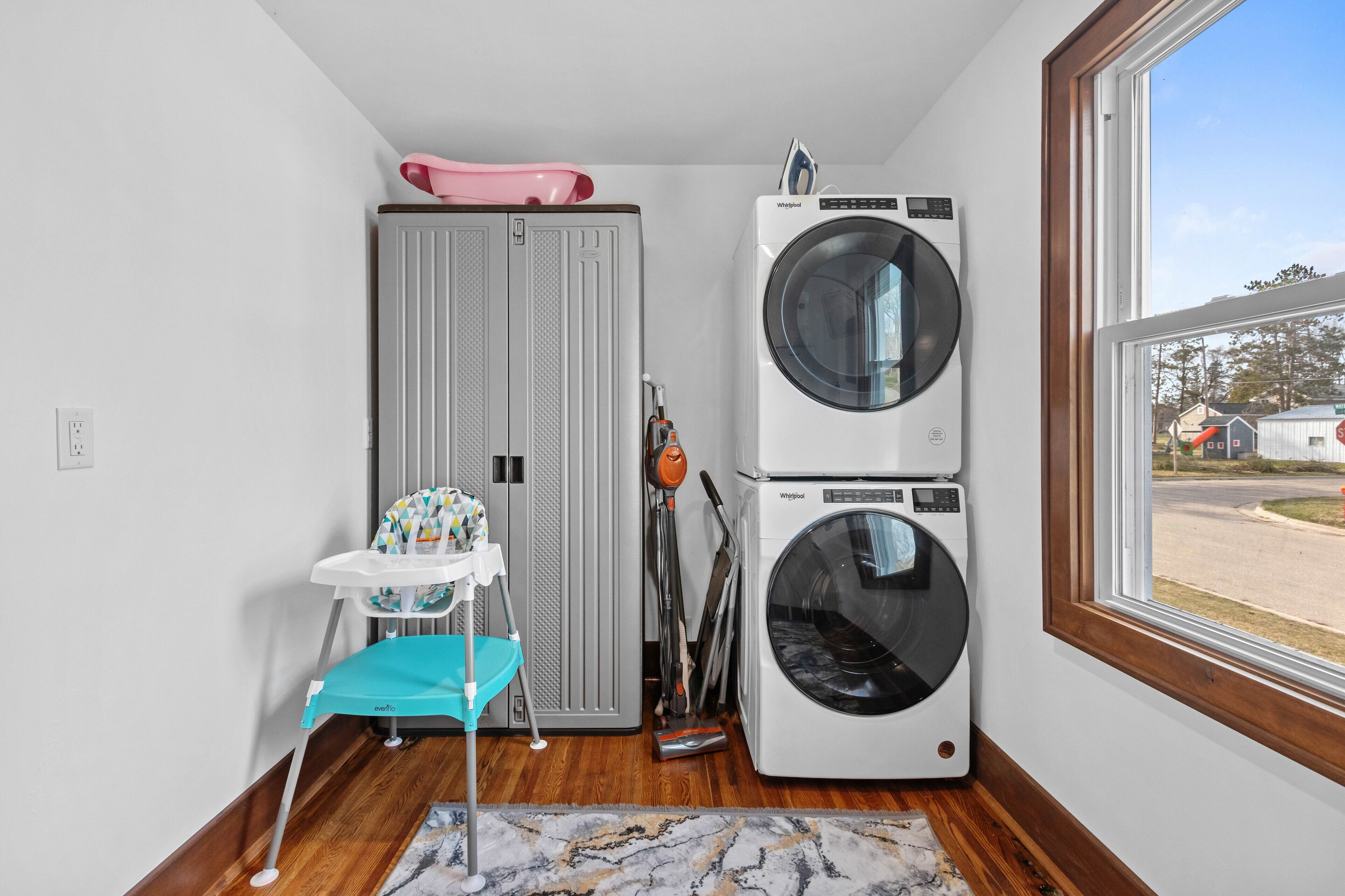 A laundry room with two stacked washers and dryers, a child’s high chair, and a window with a view of a parking lot.