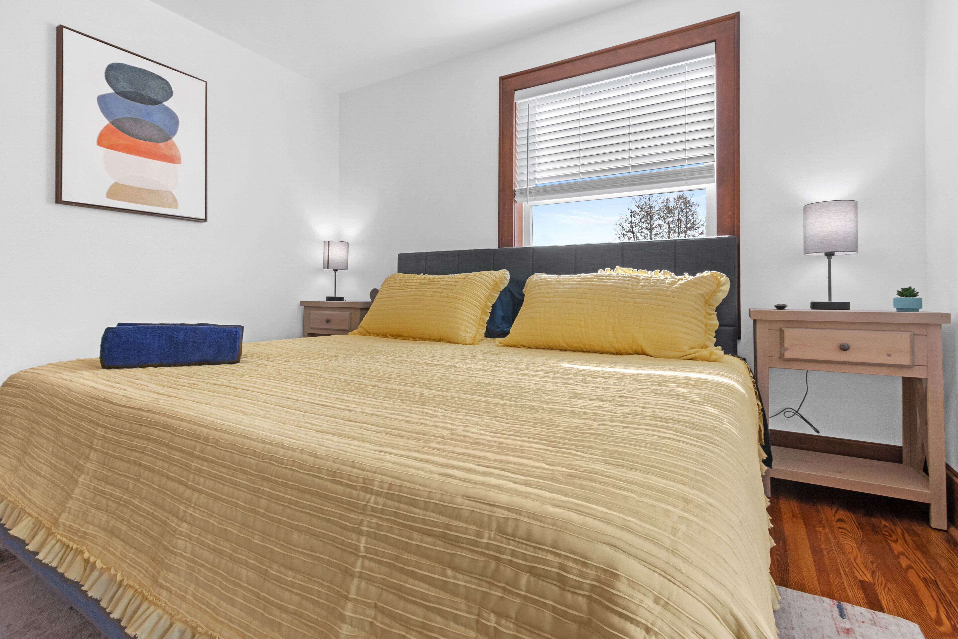 Une chambre à coucher comprenant un lit, deux lampes de chevet, une table de chevet, une fenêtre avec stores et une œuvre d’art accrochée au mur.