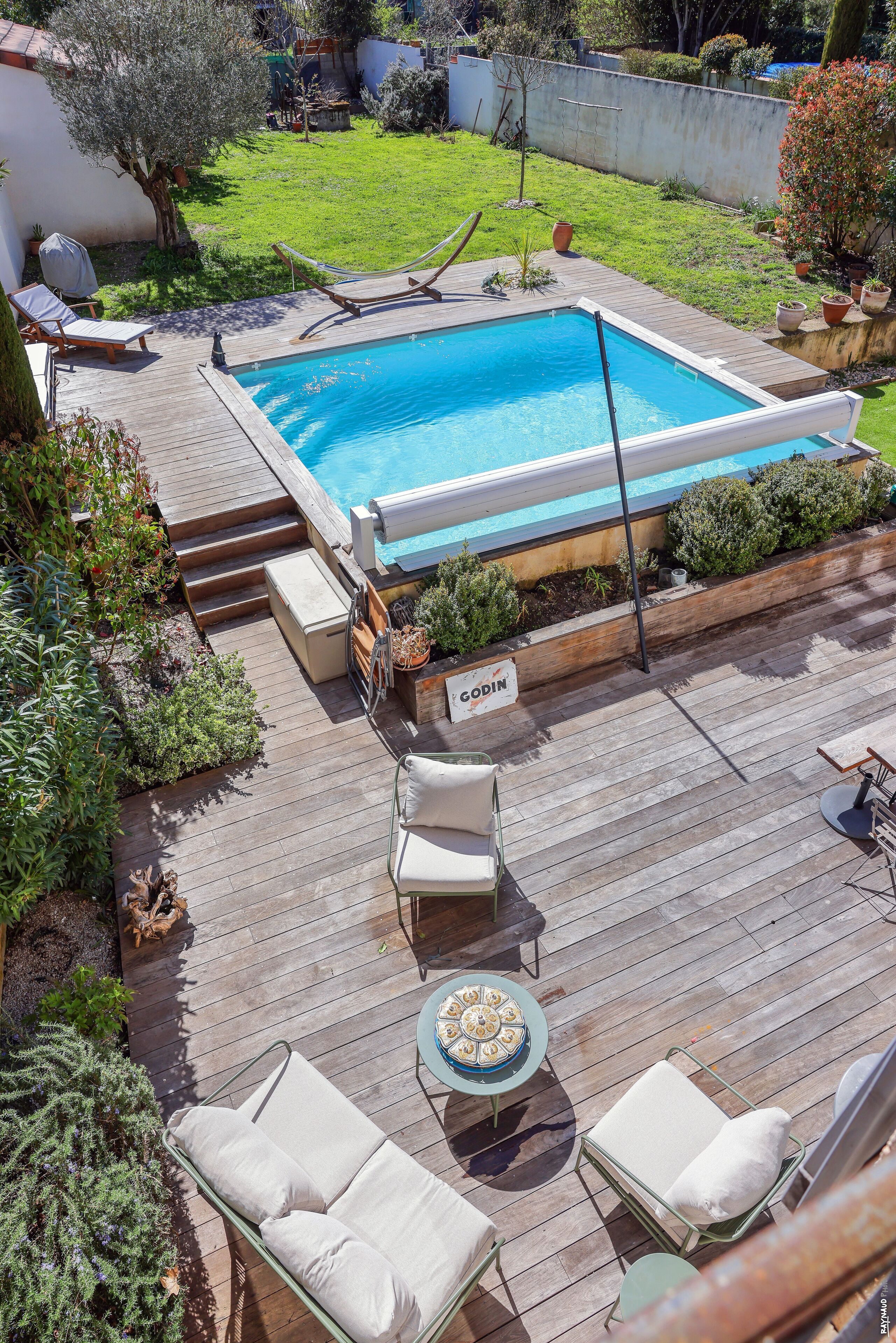 A rectangular swimming pool with a wooden deck, surrounded by potted plants and a grassy yard.
