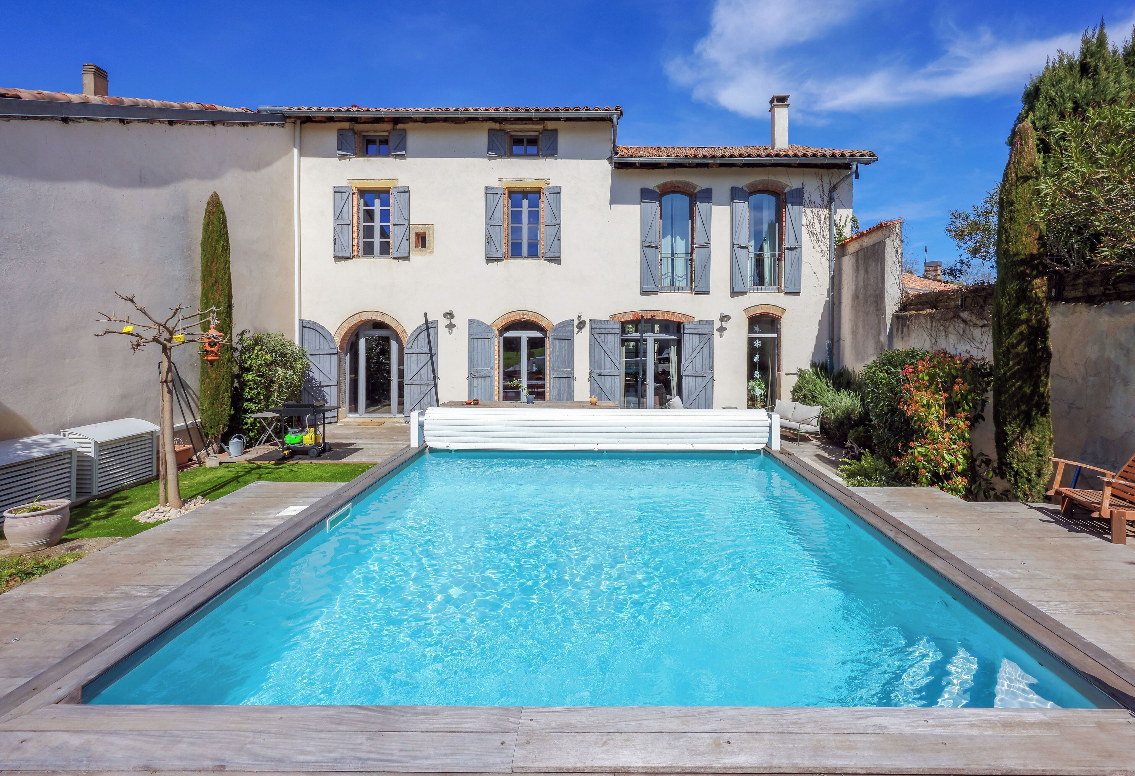 A rectangular swimming pool with a wooden deck in front, surrounded by a two-story building with arched windows and shutters.