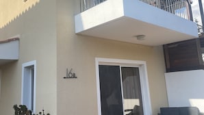 Un edificio de dos pisos con balcón, puerta corredera de vidrio y un pequeño espacio con mesas y sillas al aire libre.