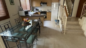 Una cocina con armarios de madera, microondas y refrigerador. Hay una mesa de comedor con sillas y superficie de vidrio. Una escalera con barandilla conduce a un nivel superior.