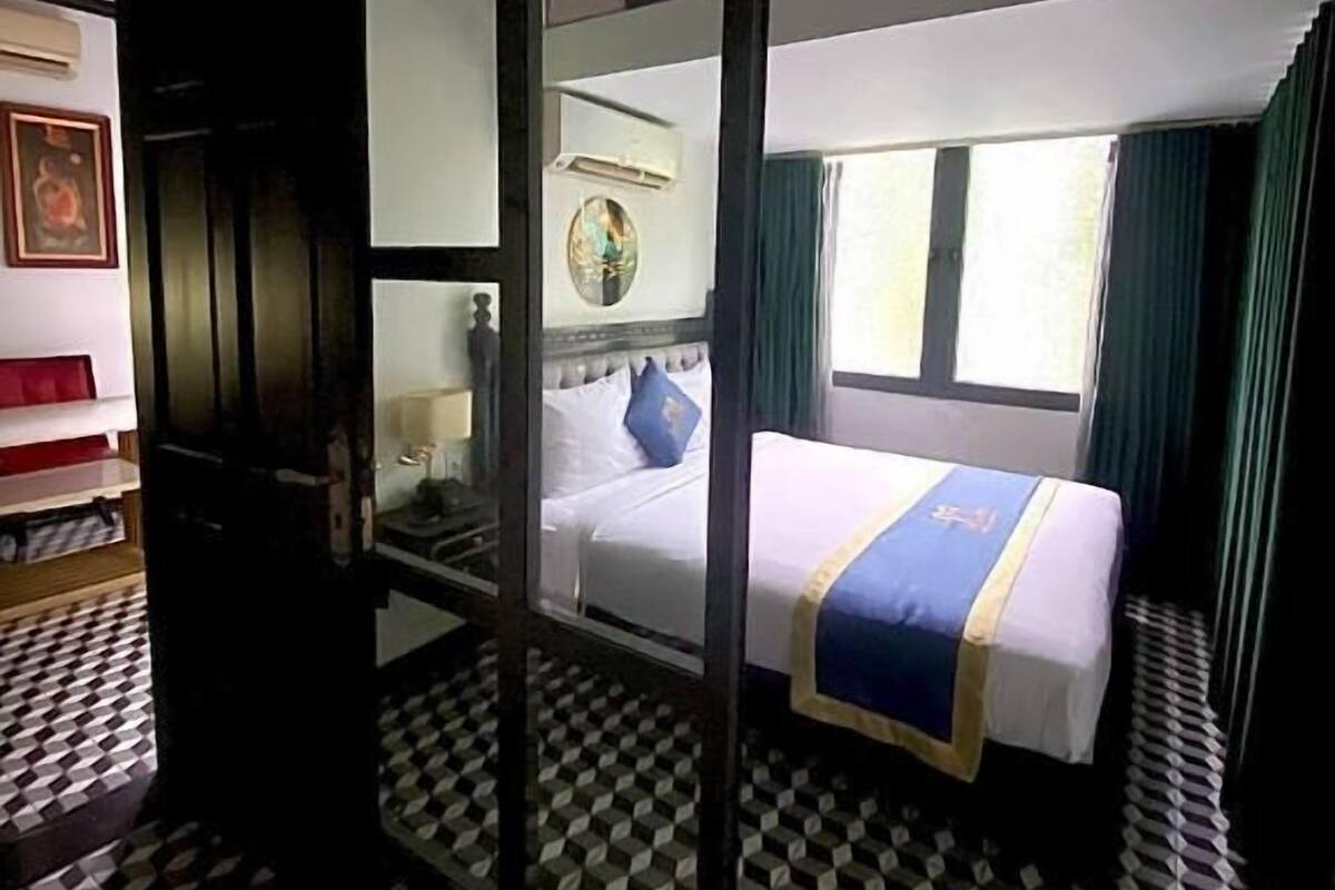 Une chambre d’hôtel avec un sol à carreaux, un lit recouvert d’une courtepointe bleue et blanche, une table de chevet, un miroir et une fenêtre avec des rideaux.