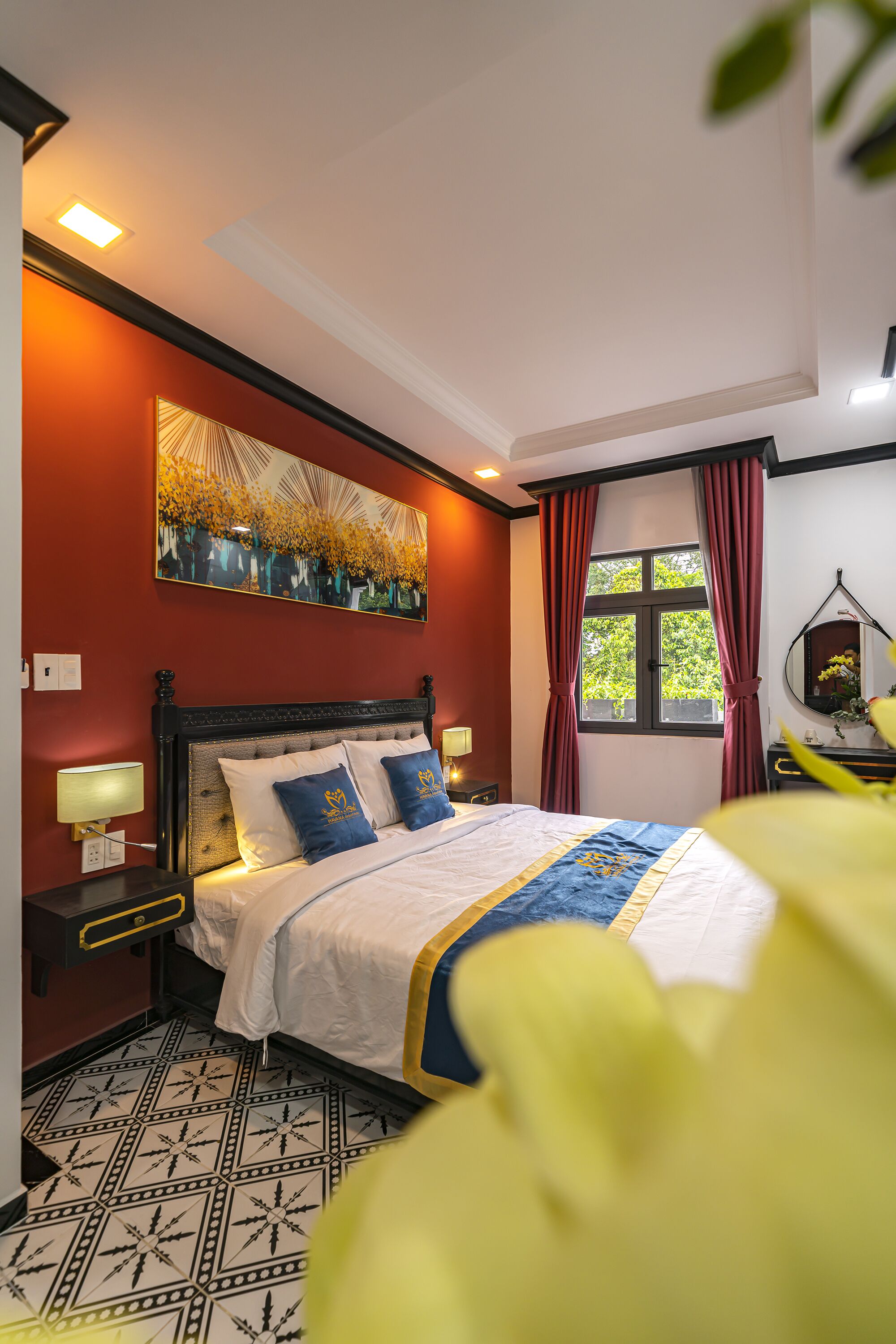 A hotel room with a bed, a nightstand, a wall-mounted lamp, a window with curtains, and a ceiling light.