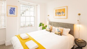 Un lit bien fait, avec du linge de lit blanc et un plaid jaune, une tête de lit grise et deux lampes fixées au mur.