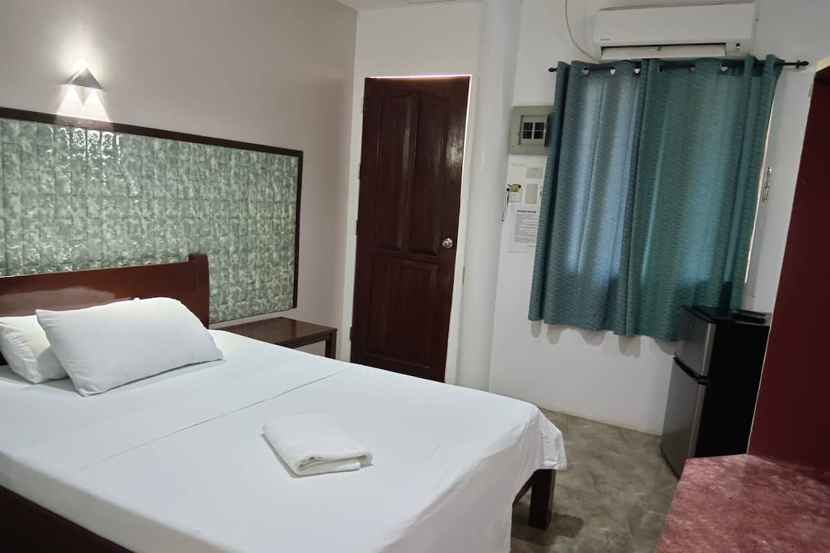 A neatly made bed with white linens and pillows, a wooden headboard, a small bedside table, a door, and a wall-mounted air conditioning unit.