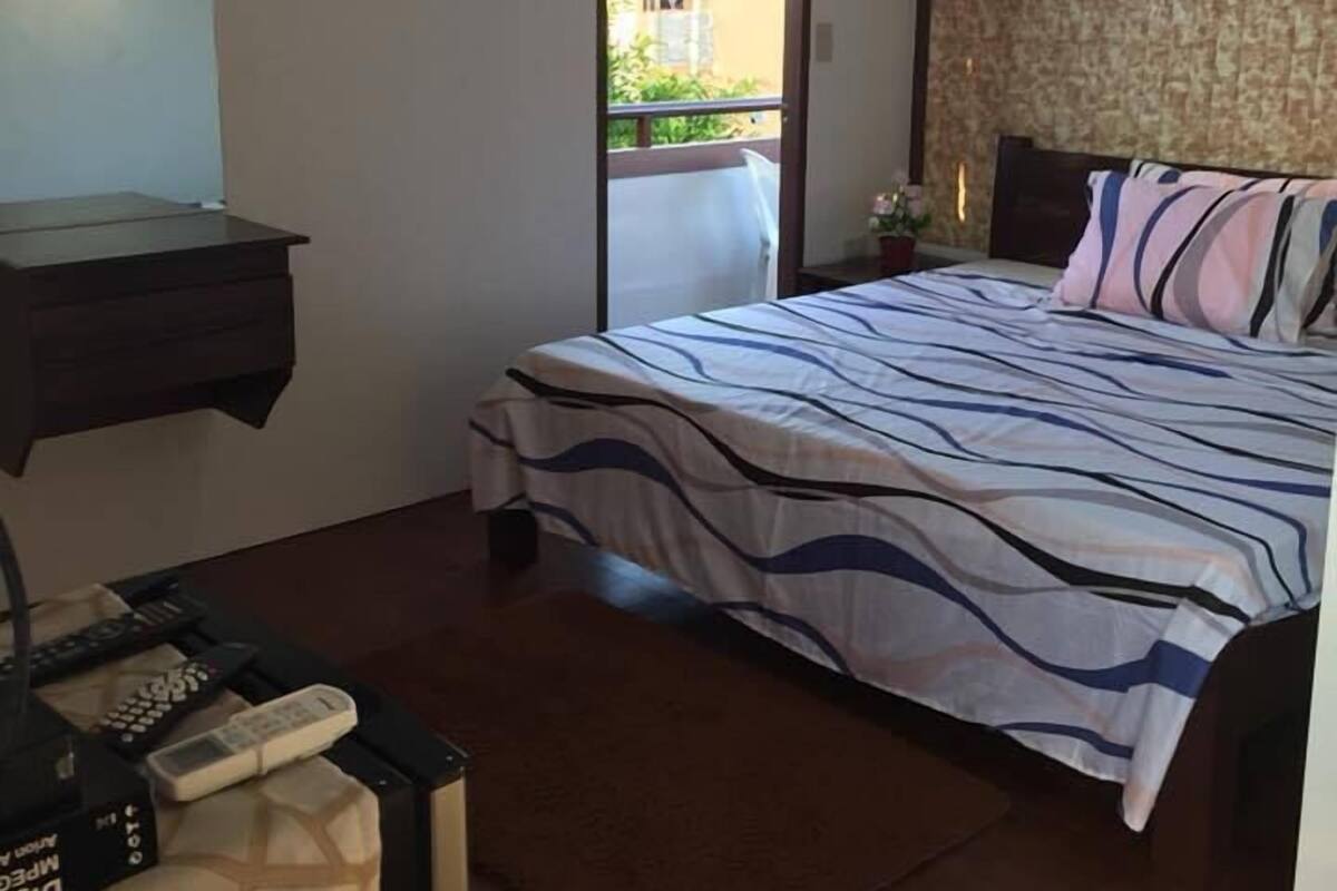 A bedroom with a bed, a desk, and a chair. There is a window with a view of buildings outside.