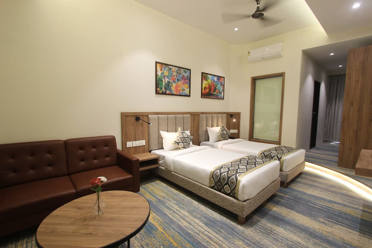 A hotel room with a brown leather sofa, a round wooden coffee table with a vase of flowers, two beds with patterned bedding, and two abstract paintings on the wall.