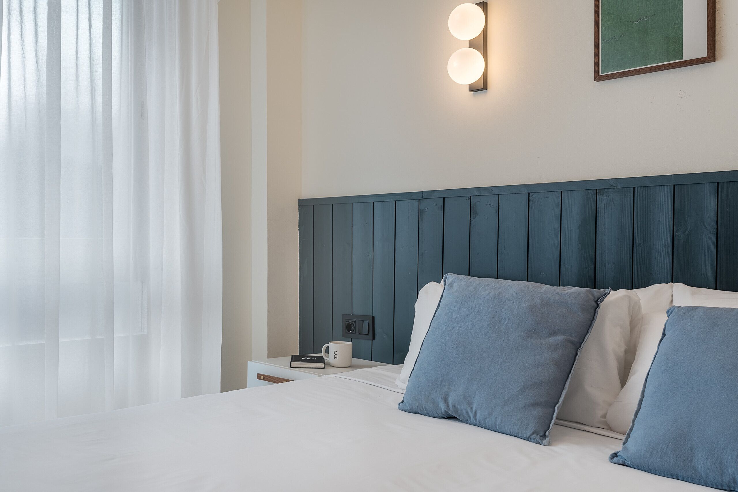 Una cama bien hecha con ropa de cama blanca y azul, una mesita de noche con un libro y una taza, y un aplique de pared.