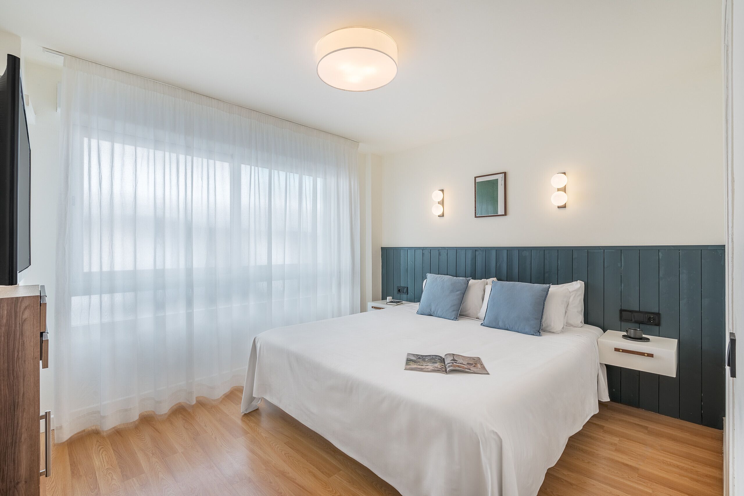 Moderni hotellihuone, jossa on suuri sänky, yöpöytä, yövalaisin ja seinään kiinnitetty valaisin.