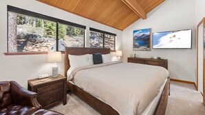 Un dormitorio con una cama grande, cabecera de madera, dos mesitas de noche, un atril, un sillón de cuero y amplias ventanas con vistas a árboles y montañas.