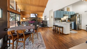 Amplia sala de estar con techo de madera, chimenea, mesa de comedor con sillas, cocina con armarios azules y un ventanal grande.