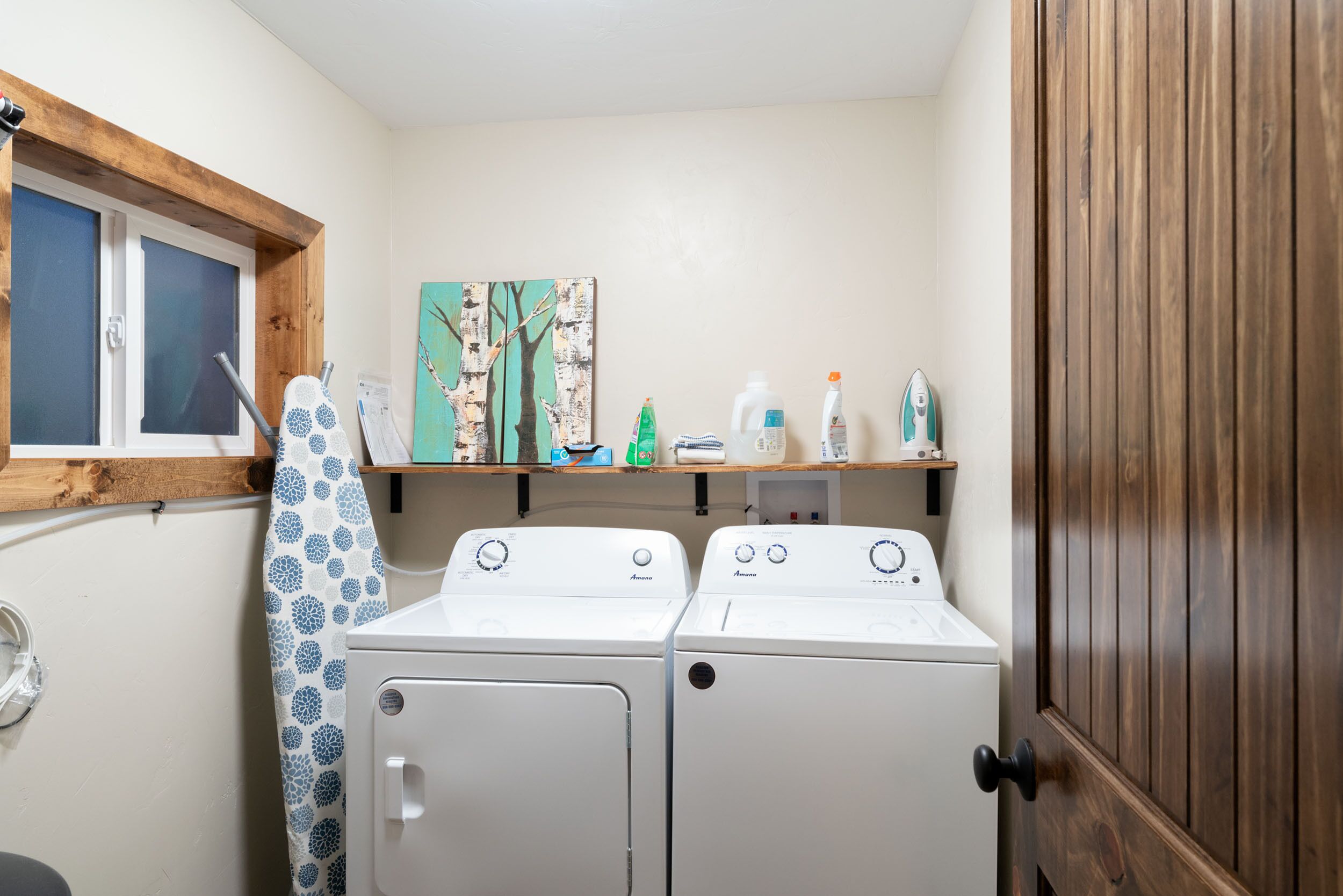 A laundry room with two front-loading washing machines, a drying rack, and a shelf with various items.