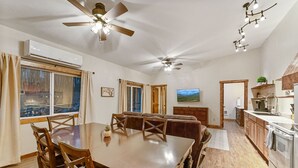 A spacious dining area with a wooden table and chairs, a ceiling fan, and a view of the outdoors through large windows. - North Fork of Spotted Bear Retreat (Columbia Falls)