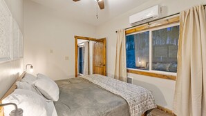 A bedroom with a bed, a ceiling fan, an air conditioning unit, a window with curtains, and a door leading to another room. - North Fork of Spotted Bear Retreat (Columbia Falls)