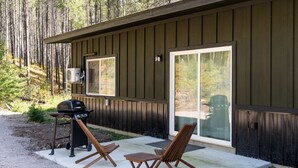 A wooden cabin with a patio area featuring a grill, two wooden chairs, and a sliding glass door. - North Fork of Spotted Bear Retreat (Columbia Falls)