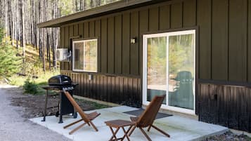 A wooden cabin with a patio area featuring a grill, two wooden chairs, and a sliding glass door.