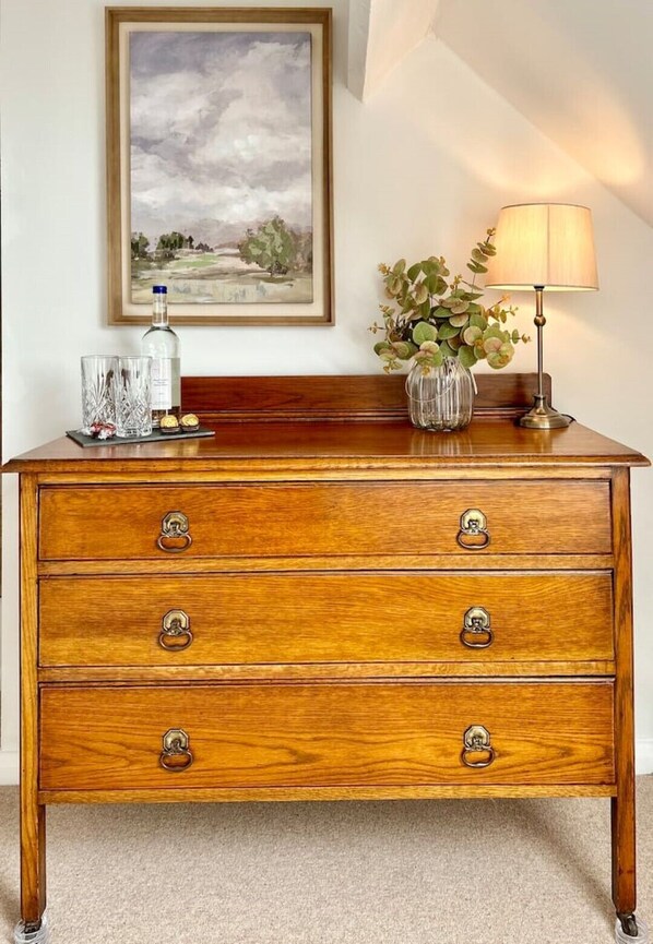 A wooden dresser with four drawers, a framed landscape painting above it, a vase with green plants, a lamp, and a bottle of water on the dresser. - Cottage in Arundel, West Sussex<br> (Arundel)