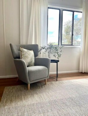 A grey armchair with a cushion, positioned on a textured rug, next to a small black side table with a plant. - Wattle Cottage (Mandurama)