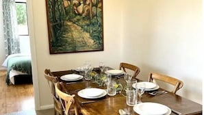 A dining table set for six with wooden chairs, a painting above it, and a view of a bedroom in the background. - Wattle Cottage (Mandurama)