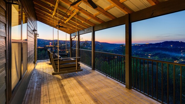 Une terrasse en bois avec une balançoire suspendue, un ventilateur de plafond et une vue sur une vallée au crépuscule.