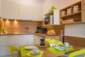 A compact kitchen with white cabinets, a microwave, and a coffee machine. There are wooden shelves above the counter holding containers and a basket of fruit.