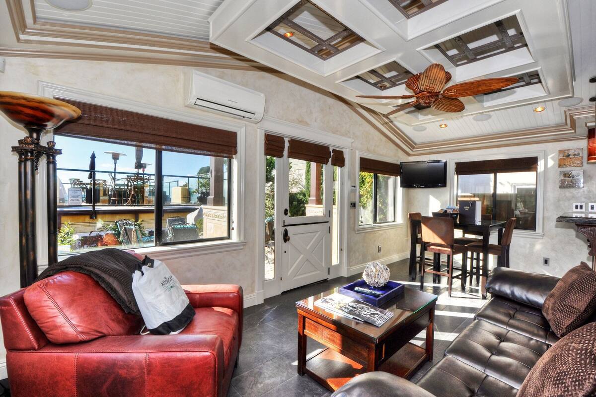 A spacious living room with a red leather sofa, a wooden coffee table, and a brown leather couch. There is a ceiling fan, large windows with blinds, and a television on the wall.