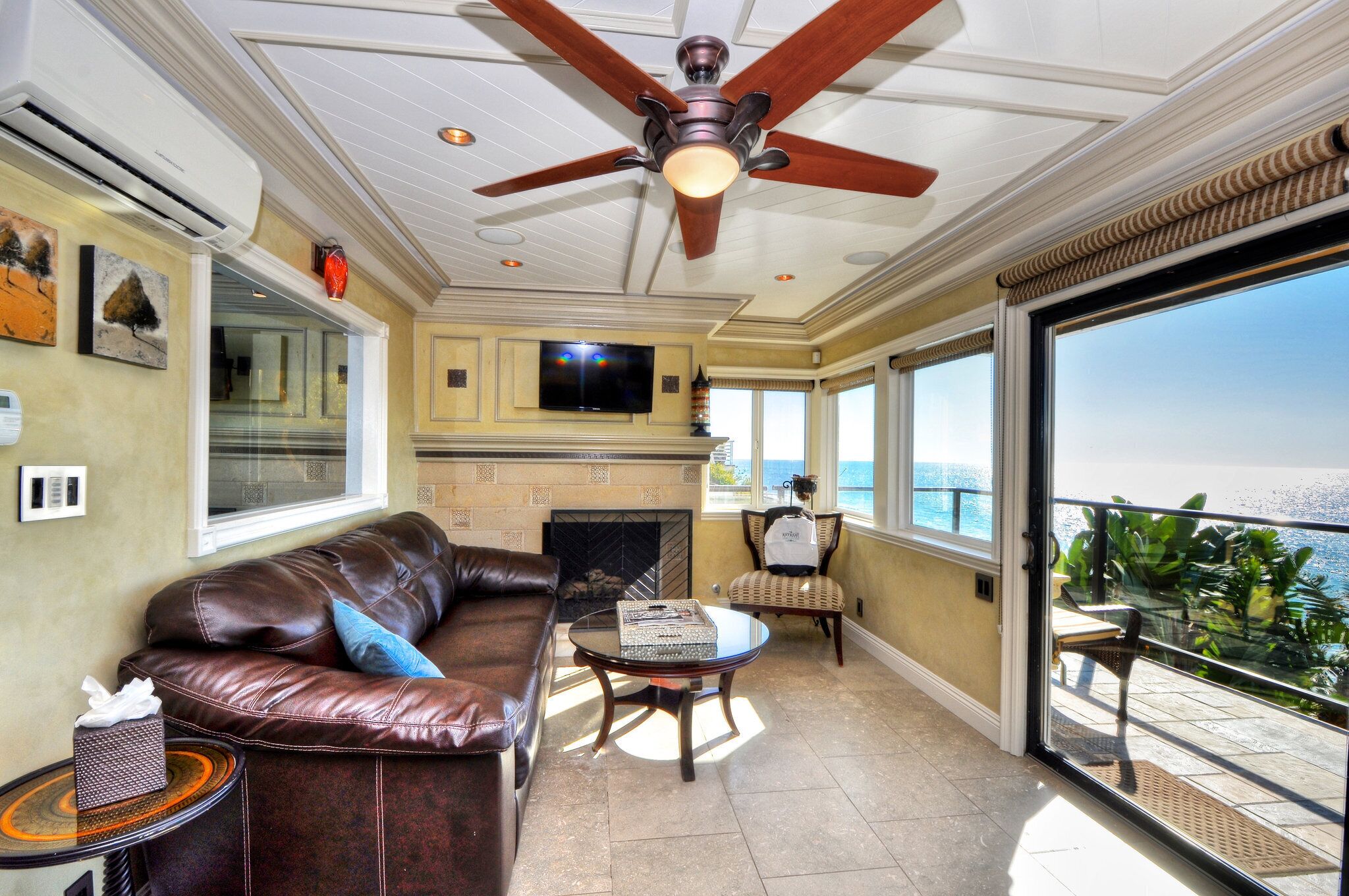 A spacious living room with a leather sofa, a glass coffee table, and a fireplace. There is a ceiling fan, a television mounted on the wall, and a balcony with a view of the ocean.