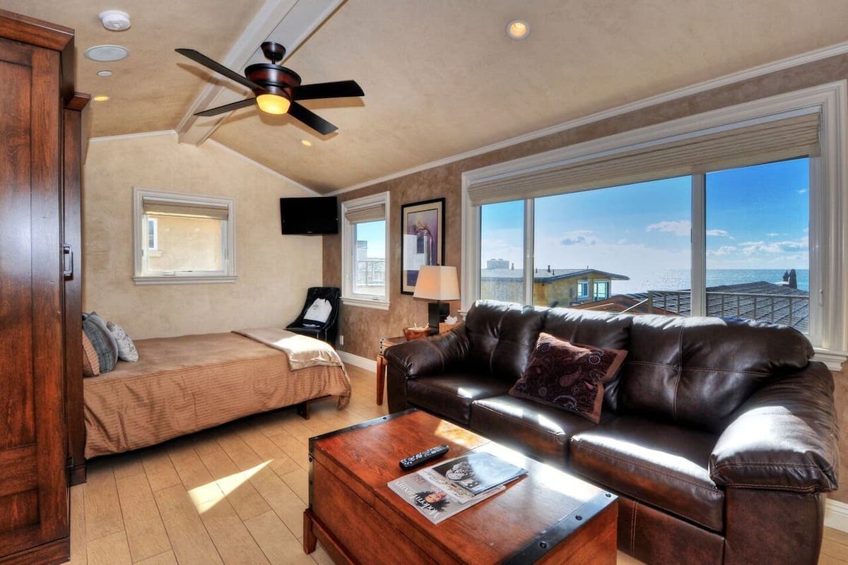A spacious living room with a leather sofa, a wooden coffee table, and a ceiling fan. There is a bed with a window, a TV mounted on the wall, and a large window offering a view of the ocean.