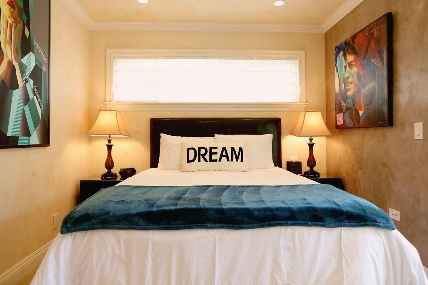 A hotel room with a bed, two bedside lamps, a window with blinds, and artwork on the walls.