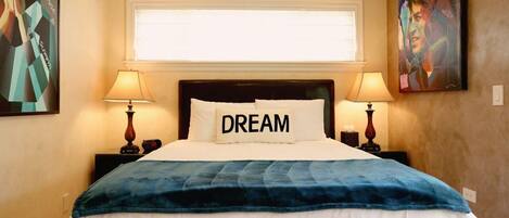 A hotel room with a bed, two bedside lamps, a window with blinds, and artwork on the walls.