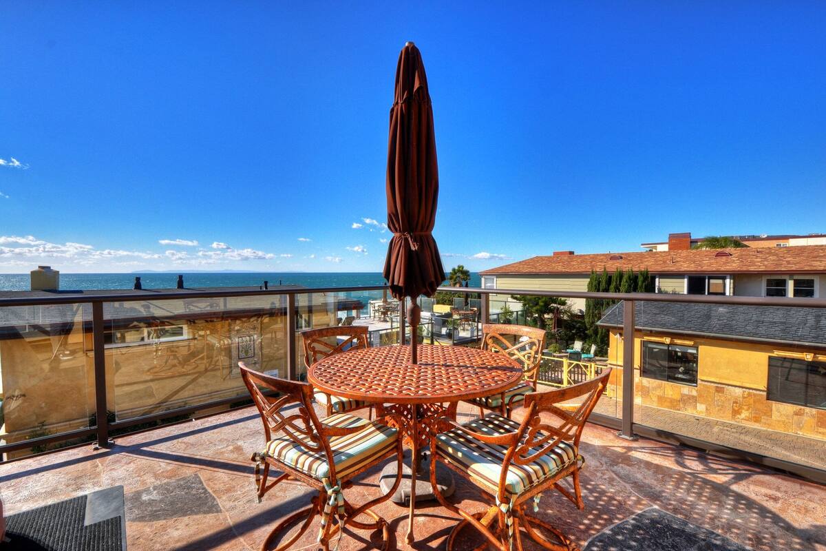 A round metal table with wicker chairs and a brown umbrella on a balcony with a view of the sea and buildings.