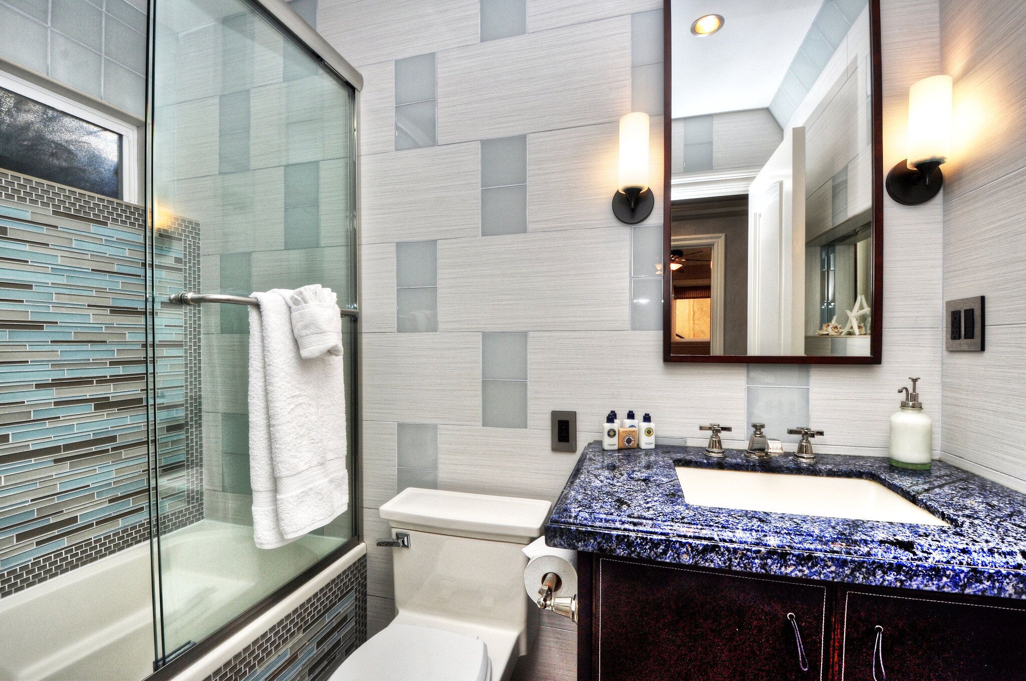 Un baño moderno con cabina de ducha de vidrio, inodoro blanco, lavabo con encimera de granito azul e iluminación empotrada en la pared sobre el espejo.