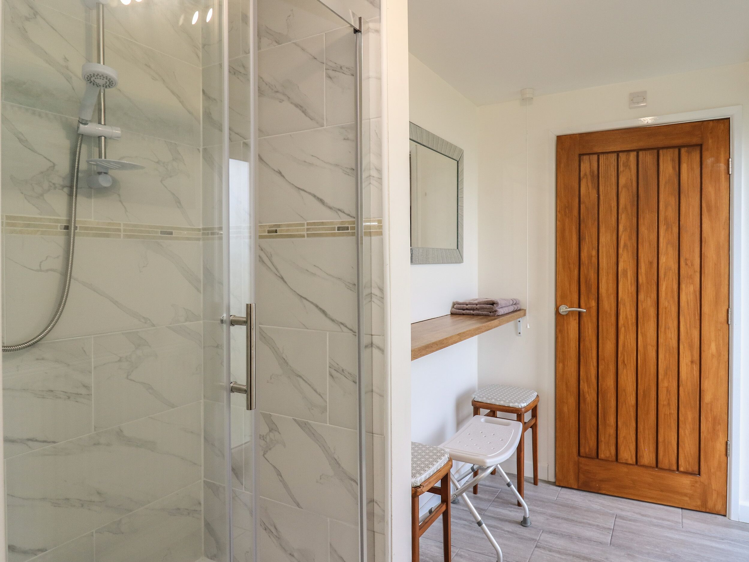 A bathroom with a glass shower enclosure, a wooden door, a small wooden shelf, and a white chair.
