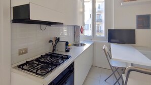 Eine kleine Küche mit weißen Schränken, einem Gasherd, einem Spülbecken und einem Fenster mit Blick auf einen Balkon.