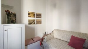 Ein Wohnzimmer mit einer Couch, einem kleinen Tisch mit einem Blumenstrauß und einem Schrank. An der Wand hängen vier gerahmte Bilder.