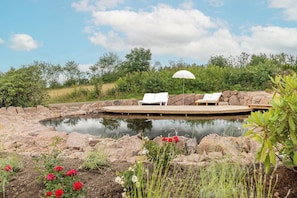 A serene outdoor pool area with lounge chairs and a wooden deck, surrounded by natural vegetation and a clear sky.